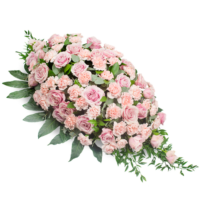 This image shows a lush, teardrop-shaped funeral spray laid out on a clean white surface, its composition carefully balanced from the full, rounded top left to the narrow, tapering tip at the lower right. The arrangement is dominated by numerous soft pink roses, their petals unfolding in delicate spirals that create a sense of depth and luxury. Some roses are fully open, with wide, layered faces that seem to cradle the light; others remain more closed, like quiet symbols of unspoken memories. Threaded between them are light peach-pink carnations, each bloom marked by characteristic ruffled petals that introduce a gentle, feathery texture. Together, the roses and carnations form a continuous blanket of pastel blossoms, creating a soothing interplay of blush and warm pink tones. Around and beneath the flowers, dense foliage anchors the spray. Broad, dark green leaves form a protective base along the outer edge, while lighter, more slender sprigs of greenery weave through the centre and trail at the pointed end, giving the piece a soft, natural finish rather than a rigid outline. The lighting is even and calm, highlighting the contrast between the vibrant greens and the muted, comforting pinks without any harsh shadows. This style of casket spray is often chosen by families in Whitechapel as a main tribute for the coffin or as a prominent display beside photographs and candles at a memorial service. In the quiet image, the spray conveys tenderness, respect, and the enduring warmth of shared memories, as though each petal holds a gentle message of sympathy and peace.