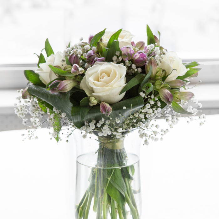 This image shows a graceful glass vase arrangement, thoughtfully designed as a sympathy bouquet and illuminated by soft, diffused daylight that gives everything a tranquil glow, much like a peaceful corner of a Whitechapel flat on a bright but gentle afternoon. At the centre, several white roses unfurl with serene precision, their creamy petals layered in smooth, overlapping curves that feel both luxurious and understated. Gathered around them are stems of alstroemeria, their blooms tinted with shades of lavender, mauve, and delicate green, some flowers open and patterned with fine markings, others still held in slender buds that hint at new blossoms yet to come. Threaded between these larger shapes are fine sprays of white baby's breath, creating a veil of tiny, airy blossoms that soften transitions and bring a sense of lightness to the composition. The flowers sit in a nest of mixed greenery, the leaves a rich, fresh green from deep, glossy ovals to narrower blades that arch gently outward, giving the bouquet structure and a natural, garden-like fullness. The clear cylindrical vase reveals the straight, green stems aligned in clear water, a simple detail that reinforces the clean, uncluttered style of the design. Behind the bouquet, the background fades into a bright, almost glowing white, with no visible objects to distract from the flowers themselves. The overall feeling is one of quiet support and heartfelt sincerity, a fitting tribute for a condolence gift or memorial service. It could easily be imagined resting on a side table in a Whitechapel home off Vallance Road or on a memory table at a small gathering near Stepney Green, its gentle palette offering wordless comfort.